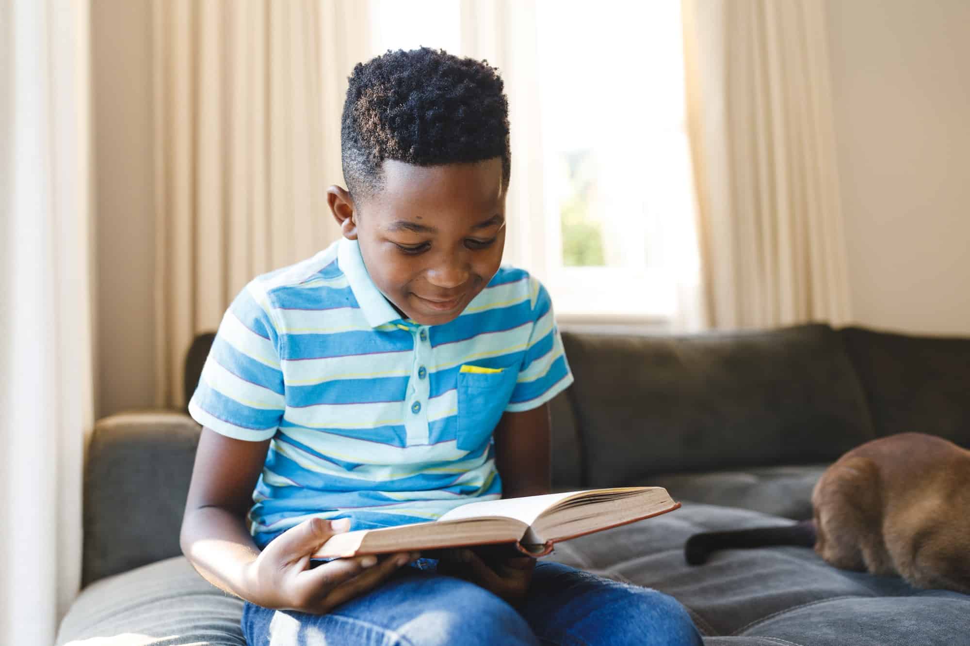 Smiling african american boy reading book and sitting on couch with cat in living room