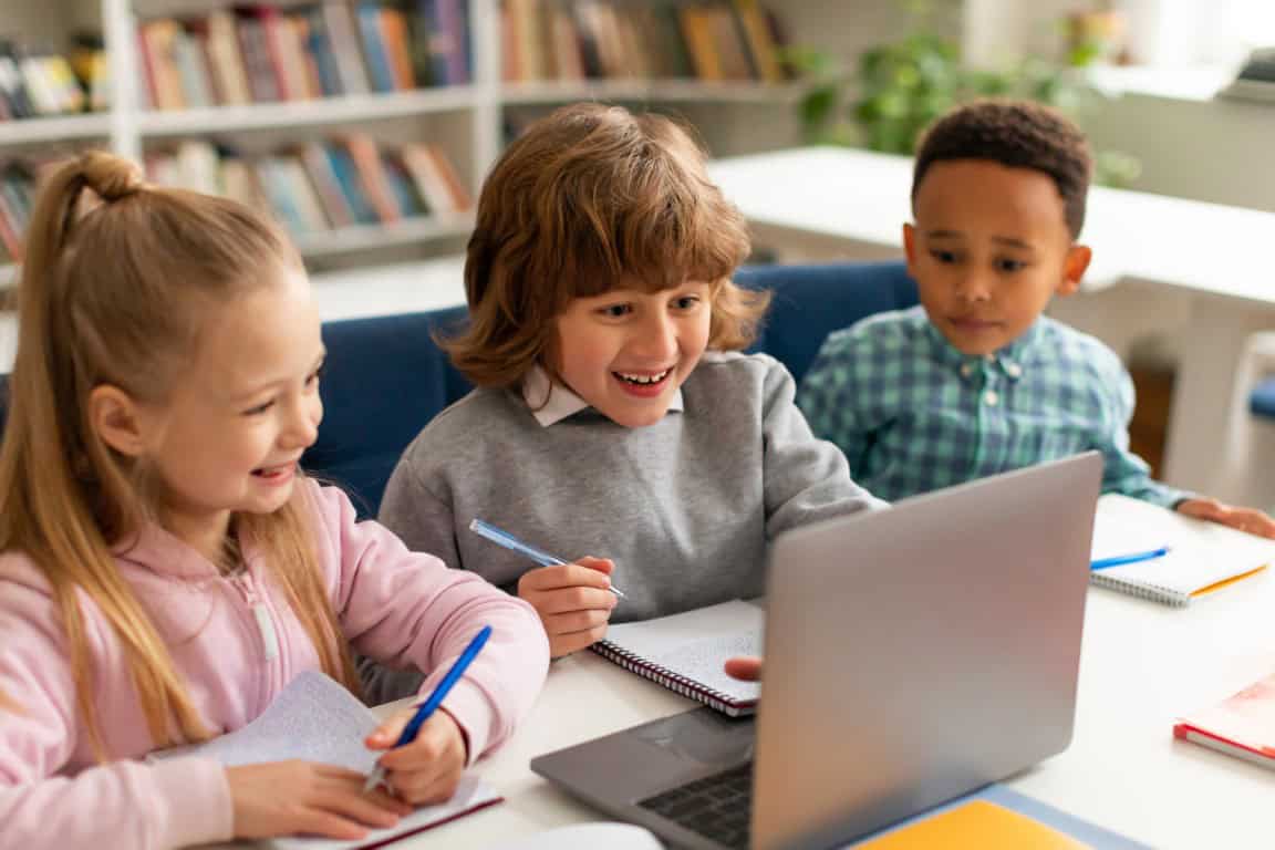 three diverse classmates sitting in front of laptop, watching videos online during lesson, looking at screen and smiling